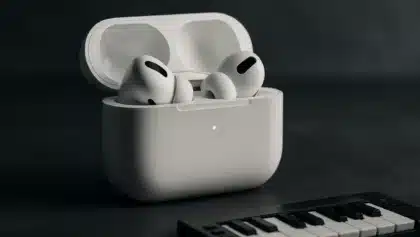 Close-up of Apple AirPods Pro in a white charging case with green LED indicator light, placed beside a keyboard.